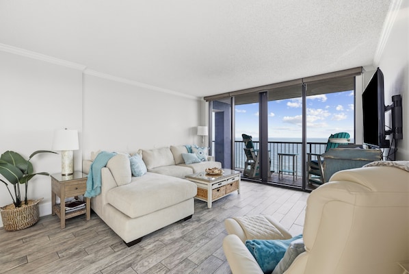 A spacious living room with a large sofa, a coffee table, and a view of the sea through a sliding glass door. - Beach Colony Oceanfront 3BR Corner Retreat (Myrtle Beach)