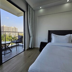 Ein Hotelzimmer mit Bett, kleinem Tisch, Stuhl, Nachttisch und einem Balkon mit Blick auf Gebäude.
