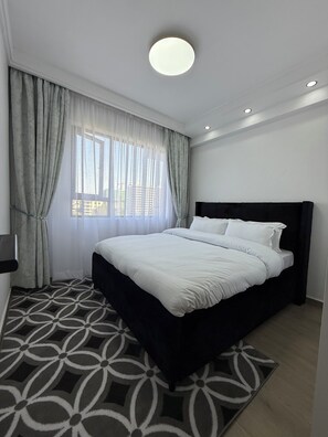 Ein modernes Schlafzimmer mit einem großen Bett, einem gemusterten Teppich und Blick auf Gebäude vor dem Fenster.