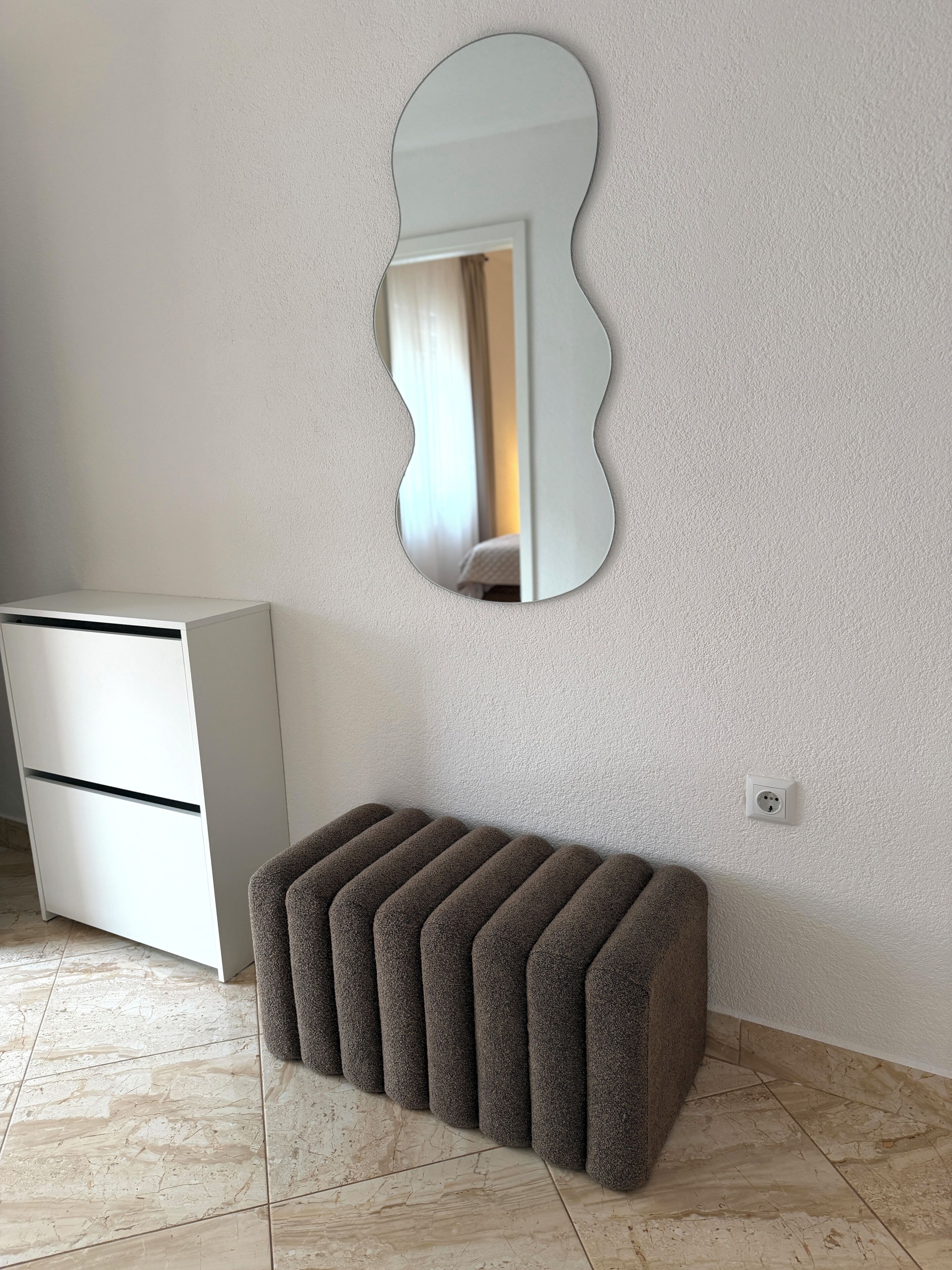 A room with a white dresser, a brown ottoman, a wall-mounted mirror, and a tiled floor.