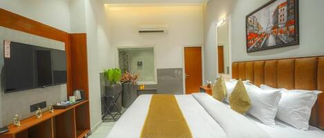 Habitación de hotel con cama, televisión, escritorio con silla, una planta y un cuadro en la pared.