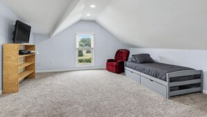Une chambre avec un plafond en pente, un lit, un fauteuil rouge, un téléviseur posé sur une étagère en bois et une fenêtre donnant sur un beau paysage.