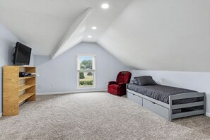 Ein Schlafzimmer mit schrägverdeckter Decke, einem Bett, einem roten Sessel, einem Fernseher auf einem Holzregal und einem Fenster mit Blick.