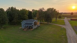 A two-story house with a deck, surrounded by trees and a dirt road. - Newly remodeled 5BR farmhouse in Coloma, just 8 minutes to Lake Michigan beach. Enjoy 3,000 sq ft, 2 full baths, smart TVs in every bedroom, a covered deck, large yard, and private balcony off the primary suite. (Coloma)