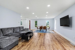 A modern living room with a sectional sofa, a dining table with chairs, and a flat-screen TV mounted on the wall. - Newly remodeled 5BR farmhouse in Coloma, just 8 minutes to Lake Michigan beach. Enjoy 3,000 sq ft, 2 full baths, smart TVs in every bedroom, a covered deck, large yard, and private balcony off the primary suite. (Coloma)