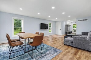 Ein modernes Wohnzimmer mit Esstisch aus Holz, braunen Stühlen, einem grauen Sofa und einem an der Wand befestigten Flachbildfernseher.
