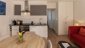 A modern kitchen with a built-in stove, a sink, and cabinets. There is a dining table with a vase of flowers. A red sofa and a lamp are visible in the living area. - Rez-de-chaussee de la Mairie - Clim - Terrasse (Arras)