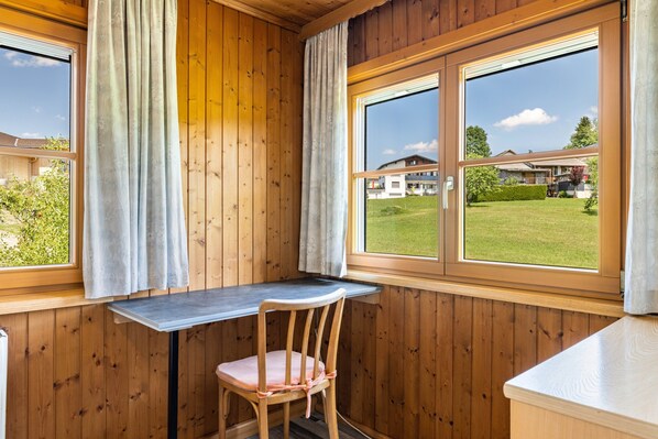 A wooden cabin room with a desk and chair, large windows, and a view of a house and lawn.