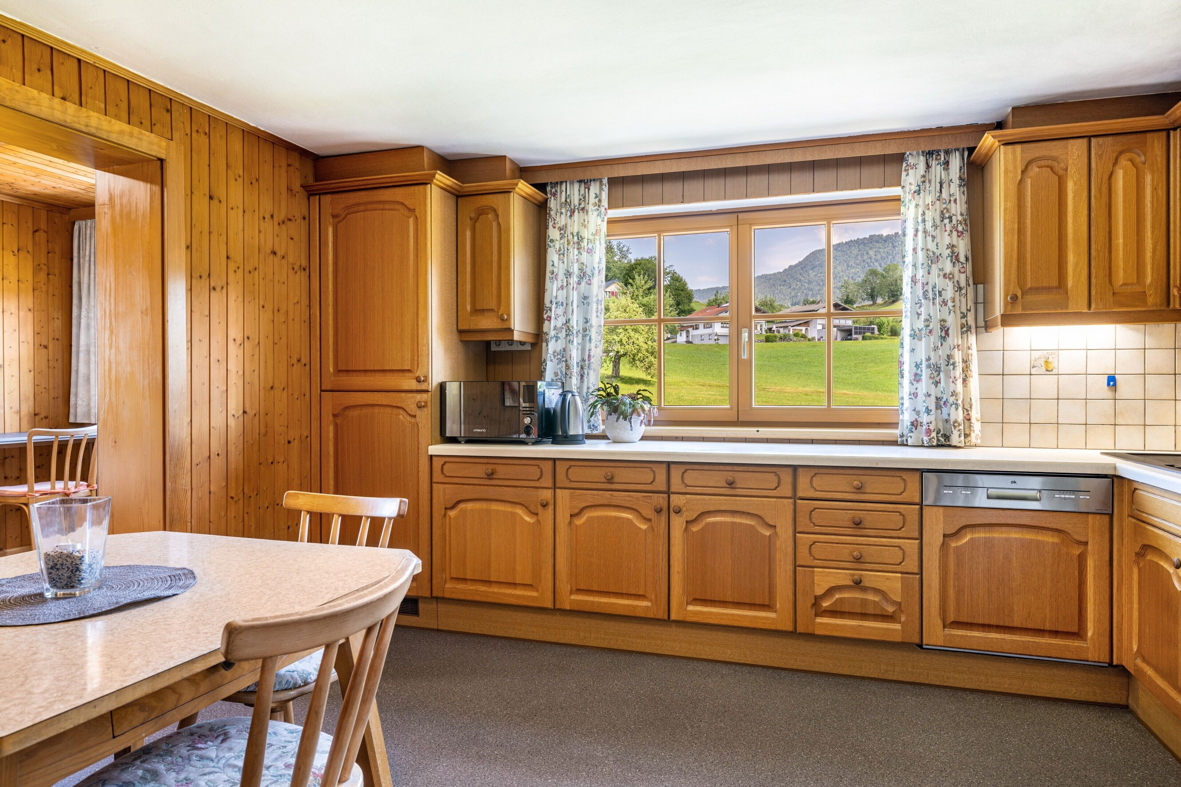 Una cocina con armarios de madera, una mesa de comedor con sillas, un ventanal con cortinas y vistas a un paisaje verde.
