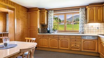 Una cocina con armarios de madera, una mesa de comedor con sillas, un ventanal con cortinas y vistas a un paisaje verde.