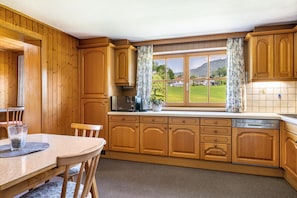 A kitchen with wooden cabinets, a dining table with chairs, a window with curtains, and a view of a green landscape.