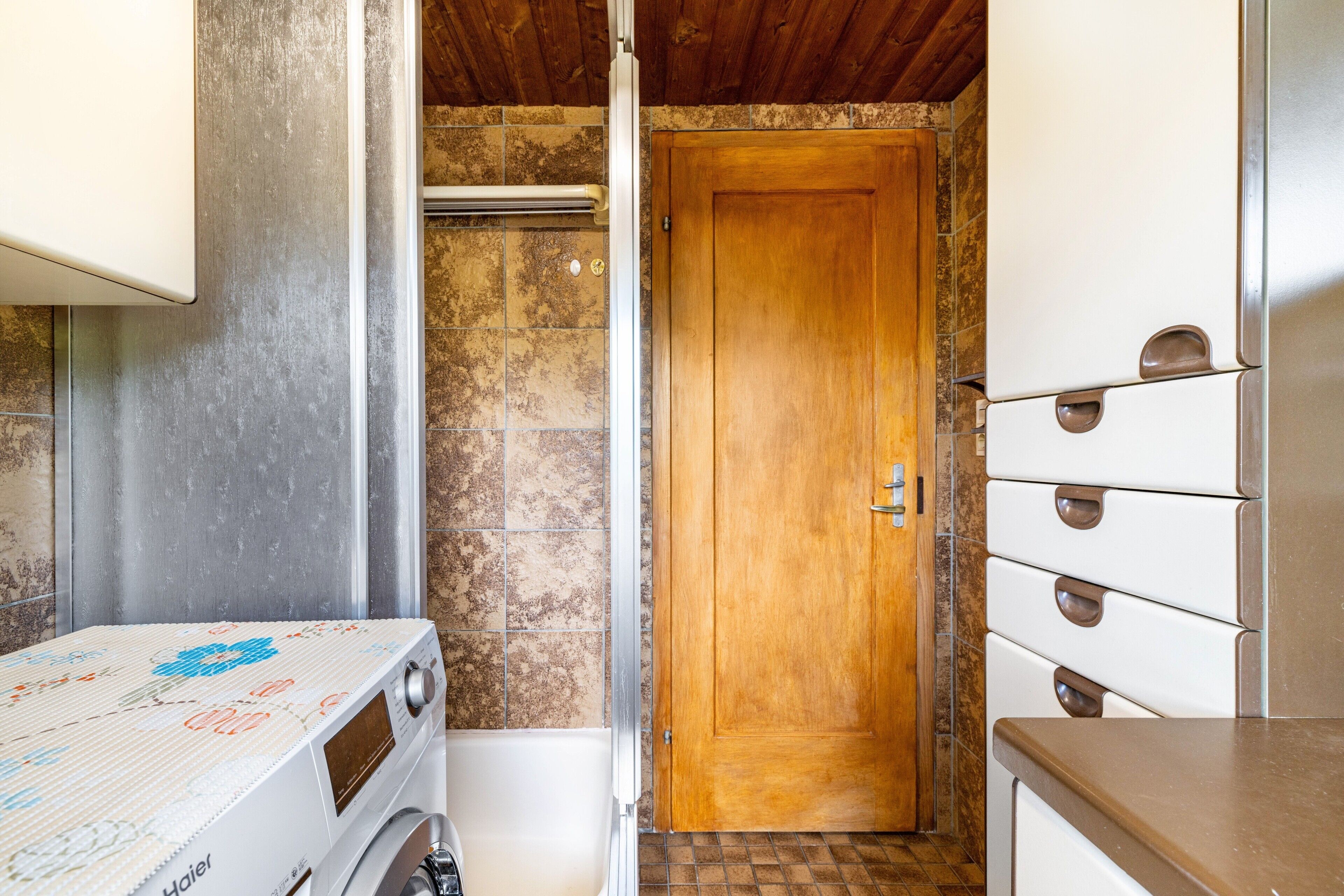 Un baño pequeño con puerta de madera, lavadora blanca y pared de azulejos.