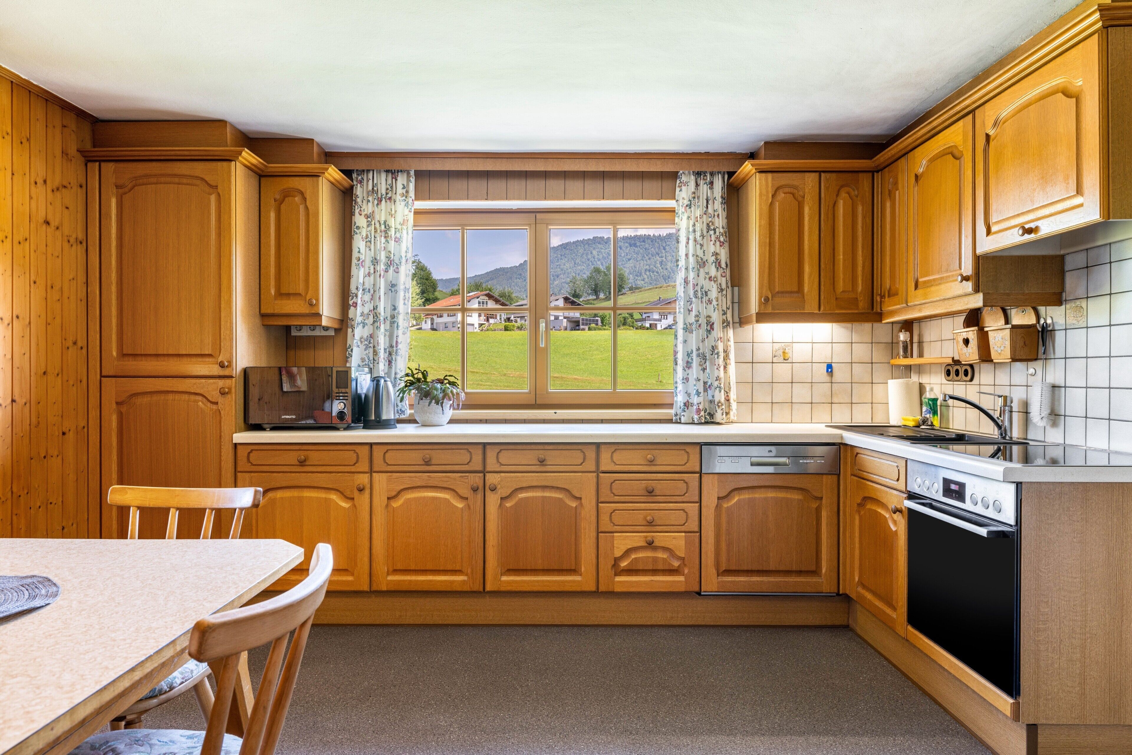 Une cuisine avec des meubles en bois, une fenêtre avec des rideaux, une table à manger et une vue sur un paysage verdoyant.