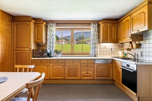 Eine Küche mit Holzausstattung, einem Fenster mit Vorhängen, einem Esstisch und Blick auf eine grüne Landschaft.