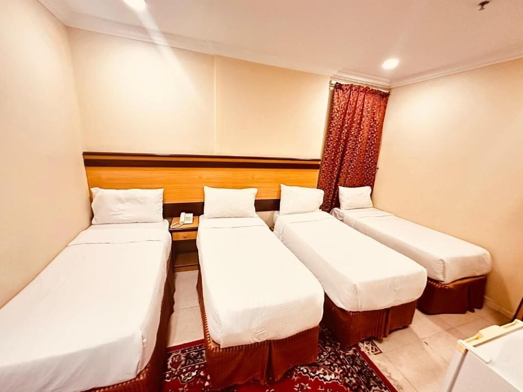 Une chambre d’hôtel avec trois lits simples alignés contre un mur, une tête de lit en bois et un rideau rouge.