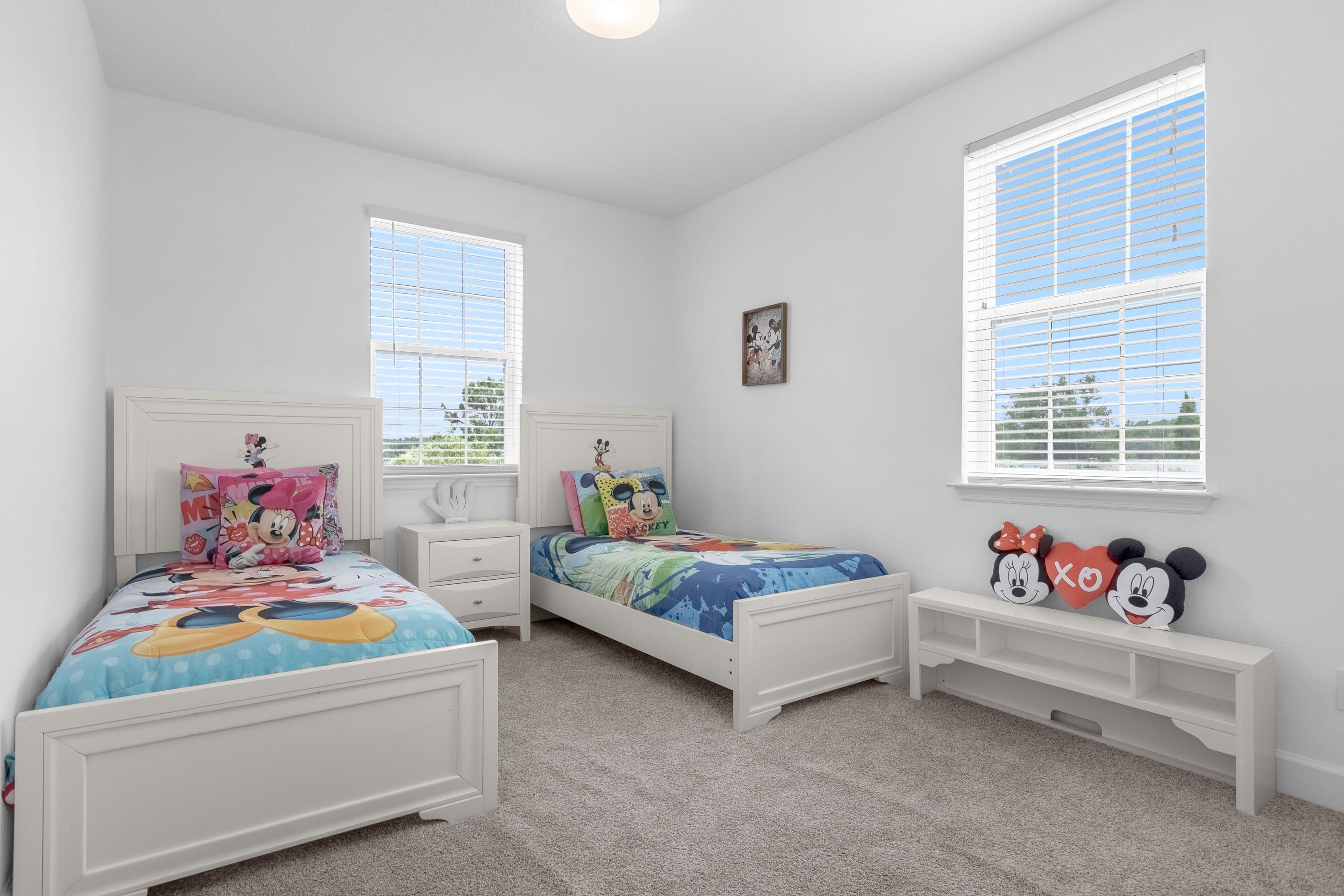 A bedroom with two beds, each with a Mickey Mouse-themed bedspread, white headboards, and a small bench between the beds.