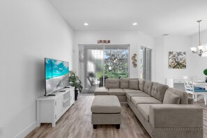 Ein modernes Wohnzimmer mit einer Eckcouch, einem Flachbildfernseher und einem Essbereich mit einem weißen Tisch und Stühlen.