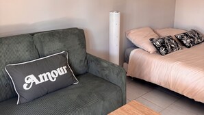 A room with a striped sofa, a bed with patterned pillows, a small table, an air conditioning unit, and a framed artwork on the wall. - Studio in the Heart of Provence (Châteaurenard)