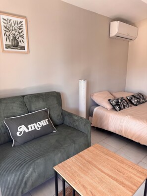 A room with a striped sofa, a bed with patterned pillows, a small table, an air conditioning unit, and a framed artwork on the wall. - Studio in the Heart of Provence (Châteaurenard)