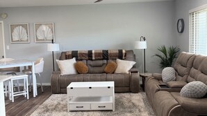 Un salon avec un ventilateur de plafond, un canapé, une table basse, un coin repas avec une table et des chaises, et une plante.