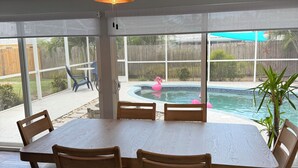 Un coin repas comprenant une table et des chaises, un ventilateur de plafond et une vue sur un jardin avec une piscine et une clôture en bois.