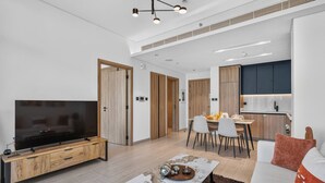 Ein modernes Wohnzimmer mit Flachbildfernseher, Holzmöbeln, einem Essbereich mit Stühlen und einer Küche mit dunklen Schränken.