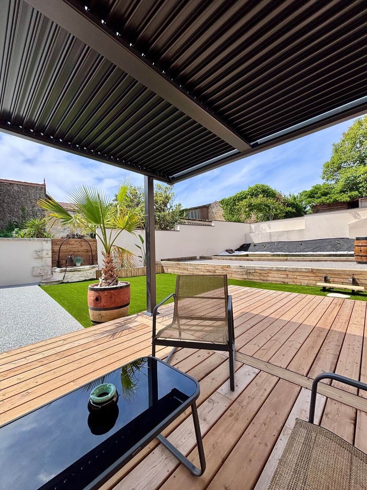 Une terrasse extérieure avec une table et des chaises, une pergola au-dessus et une vue sur un jardin et un bâtiment en arrière-plan.