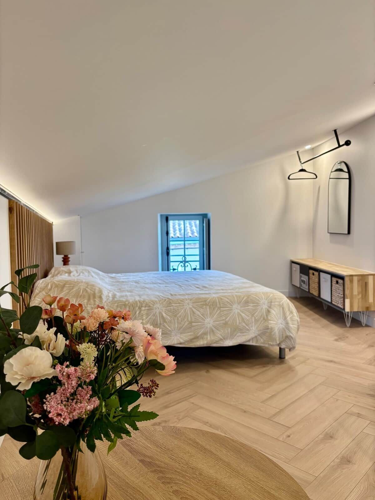 A bedroom with a bed, a window, a bedside table, a mirror, and a vase with flowers.