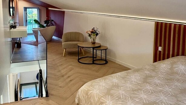 A modern bedroom with a large bed, a small round table, and a chair. There is a kitchenette area with a sink and a window.