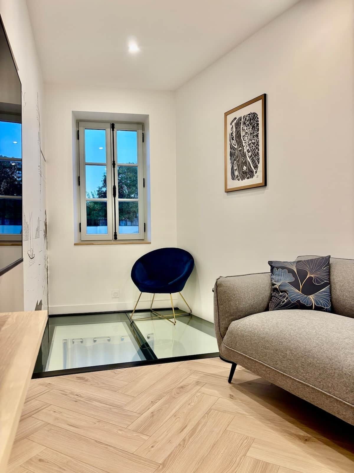 A modern living room with a glass table, a blue chair, a grey sofa, and a framed artwork on the wall.