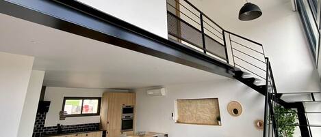 A modern interior with a grey sofa, dining table, kitchen, staircase, and large windows.