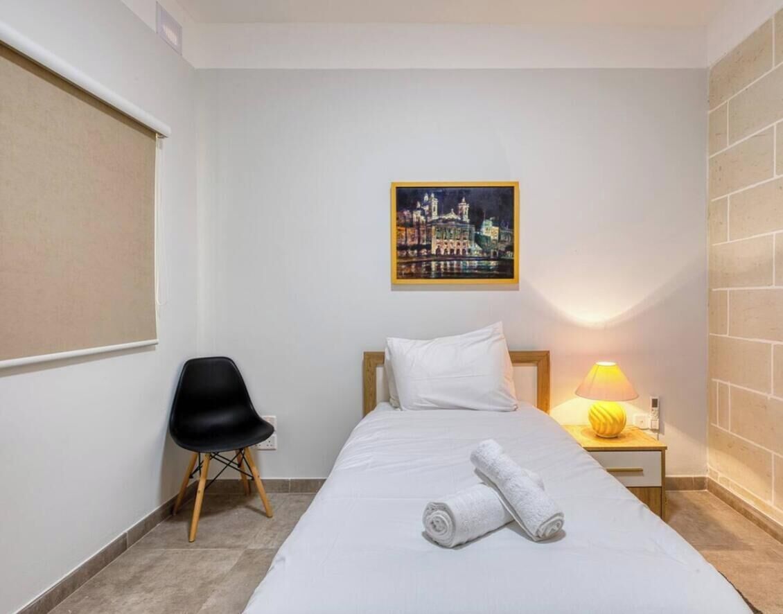A hotel room with a bed, a chair, a nightstand with a lamp, and a cityscape painting on the wall.