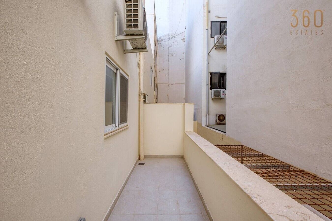 Un couloir étroit au sol carrelé, avec une fenêtre sur la gauche et des unités de climatisation fixées aux murs.