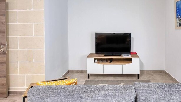 A living room with a flat-screen TV on a wooden TV stand, a sofa, and a patterned rug. - 3BR Apartment with AC & Balcony in Marsascala (Marsaskala)