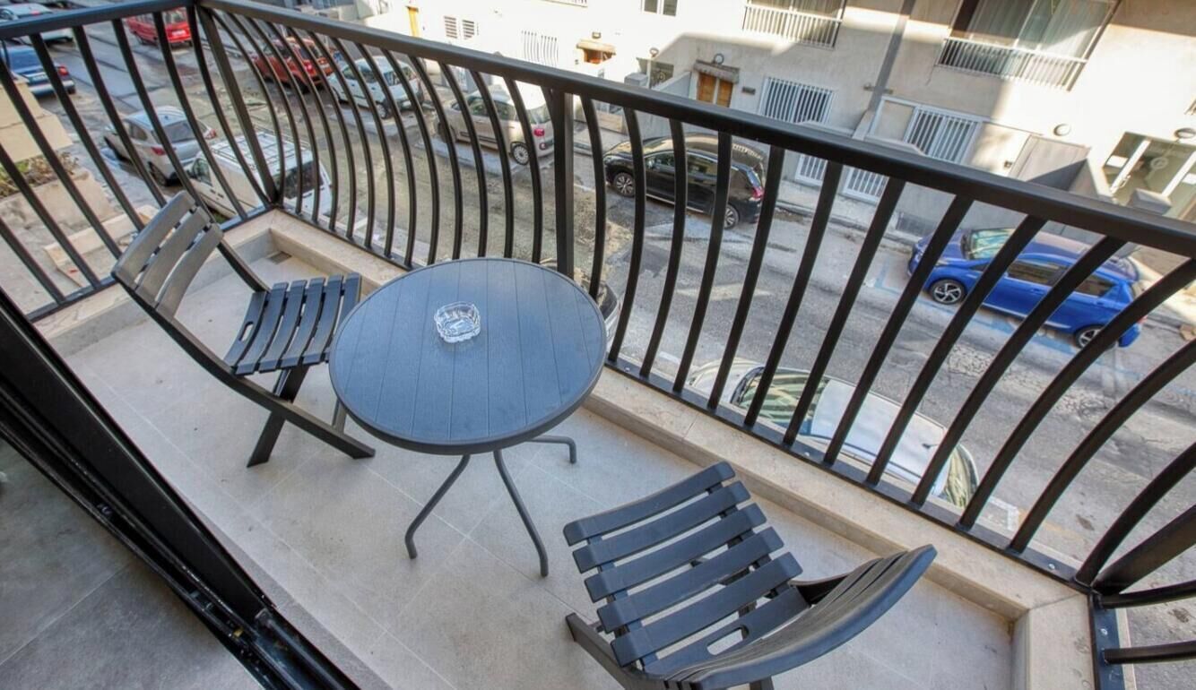 A balcony with a table and chairs, a metal railing, and a view of parked cars and buildings.