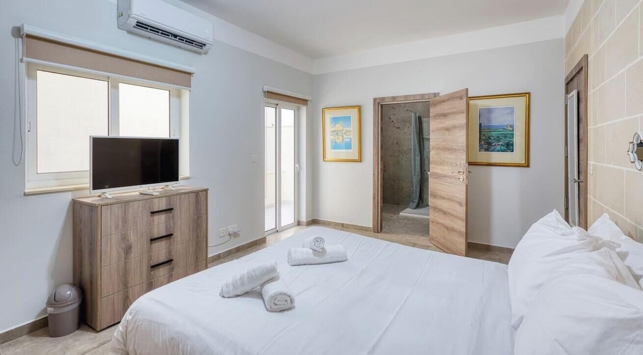 A hotel room with a bed, a TV, a wooden dresser, a window, and a door leading to a shower area.