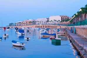 A marina with boats docked along a waterfront promenade, buildings in the background, and a clear sky. - 3BR Apartment with AC & Balcony in Marsascala (Marsaskala)