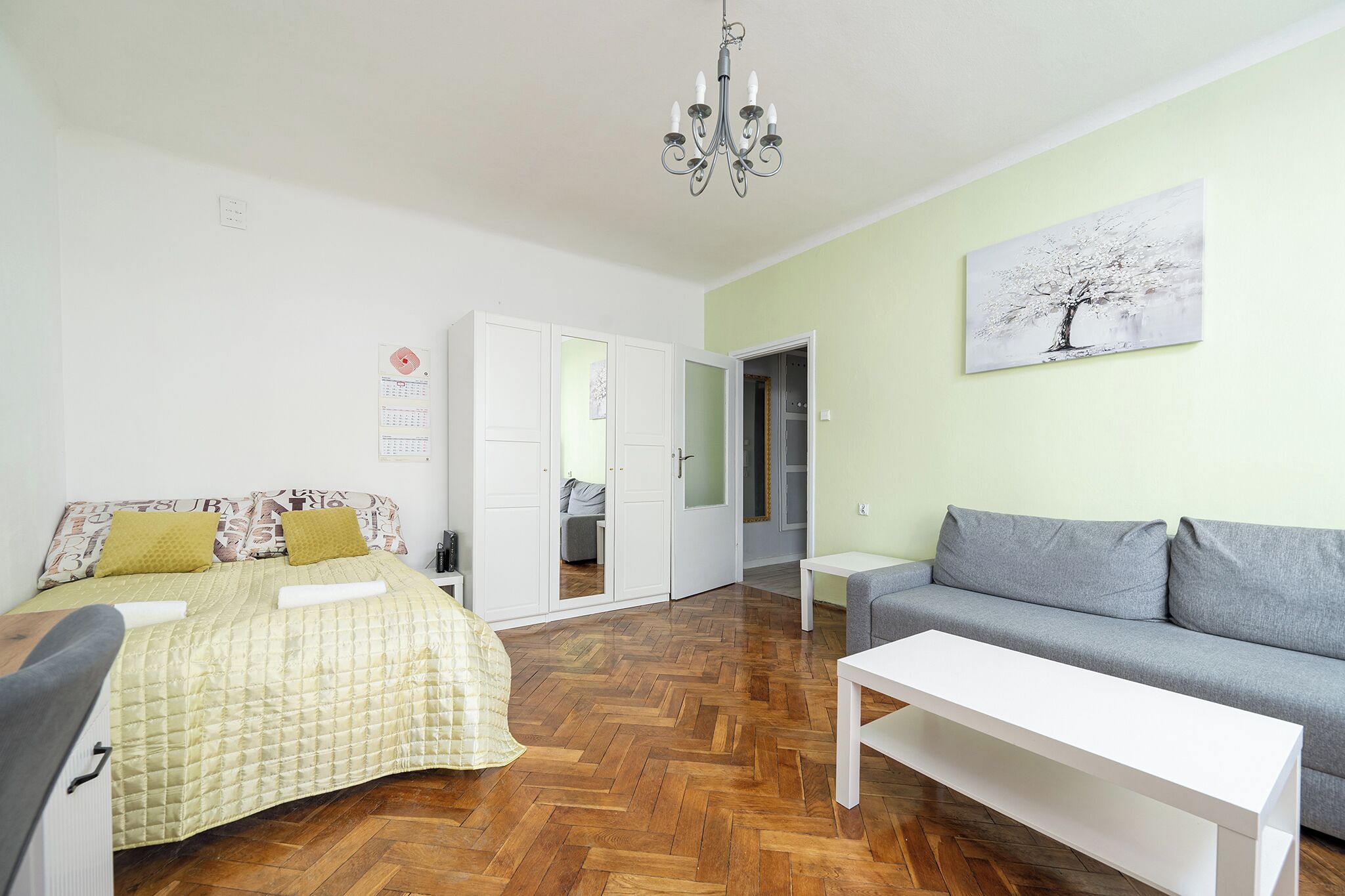 Ein Zimmer mit einem Bett, einem Schlafsofa, einem weißen Tisch, einer Chandelüster und einem Gemälde an der Wand.