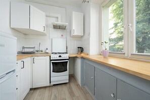 A kitchen with white cabinets, a stove, a sink, and a window with a view of trees. - RentPlanet - Apartament Orla (Warszawa)