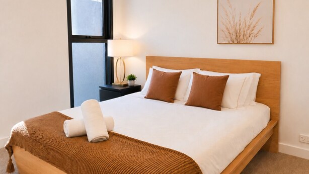 Ein ordentlich bezogenes Bett mit brauner Decke und Kissen, ein Nachttisch mit Lampe und ein gerahmtes Bild an der Wand.
