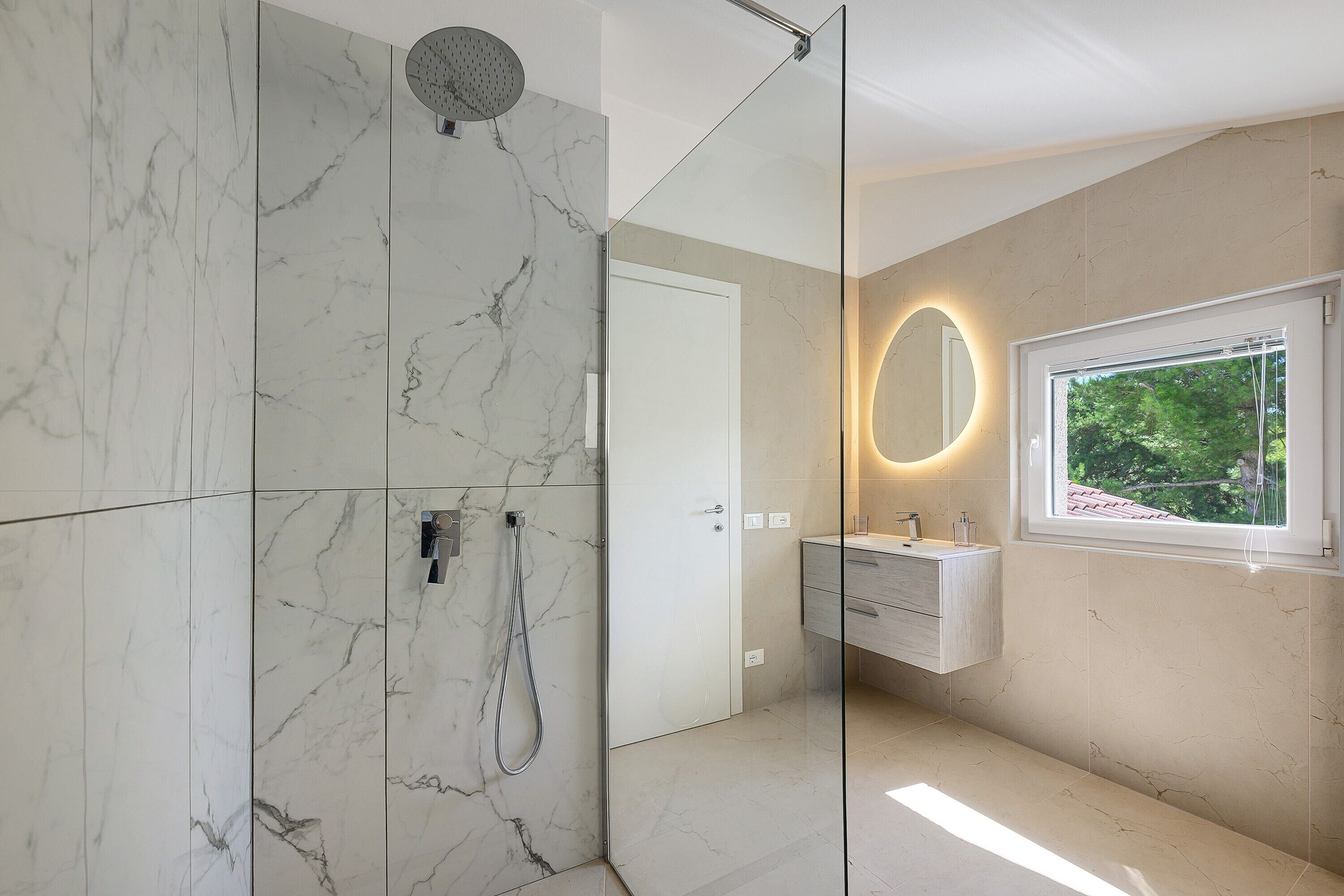 A modern bathroom with a glass-enclosed shower, a vanity with a mirror, and a window providing a view of greenery.