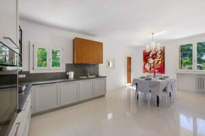 Une cuisine moderne dotée d’un îlot central, d’un coin repas agrémenté d’un lustre, et de grandes fenêtres offrant une vue sur la verdure.