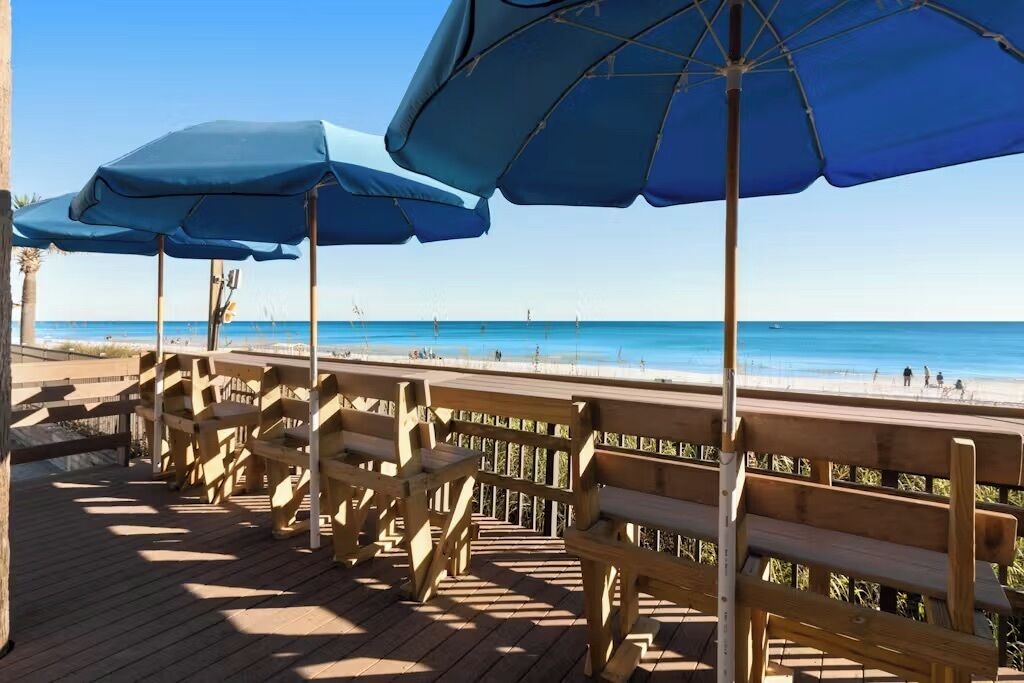 A wooden deck with benches and blue umbrellas overlooking a beach.