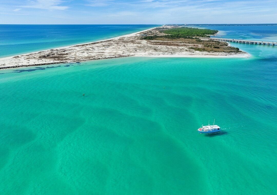 Vista aérea de una zona costera con aguas turquesas cristalinas, una pequeña isla y un muelle que se adentra en el mar.