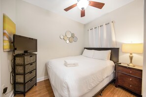 Een hotelkamer met een bed, een televisie op een standaard, een plafondventilator, een nachtkastje met een lamp en een spiegel aan de muur.