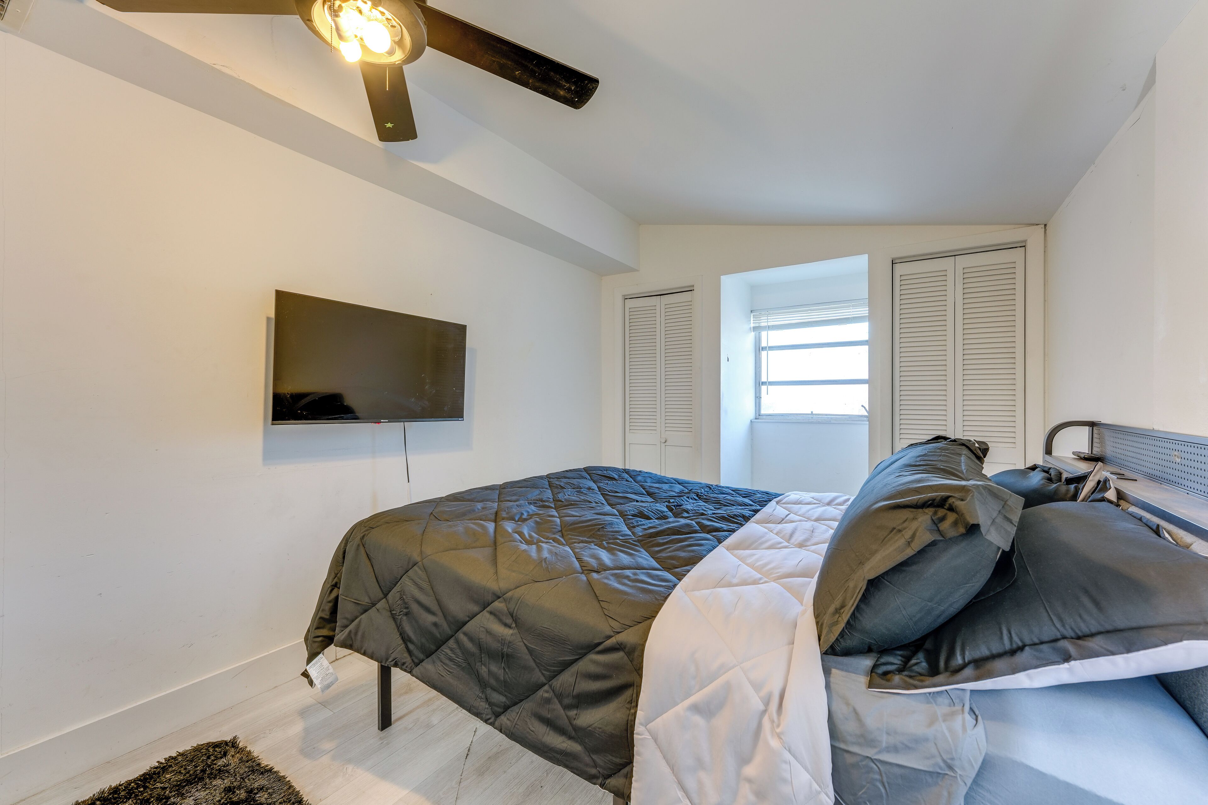 A bedroom with a bed, a ceiling fan, a mounted TV, and a window with blinds.