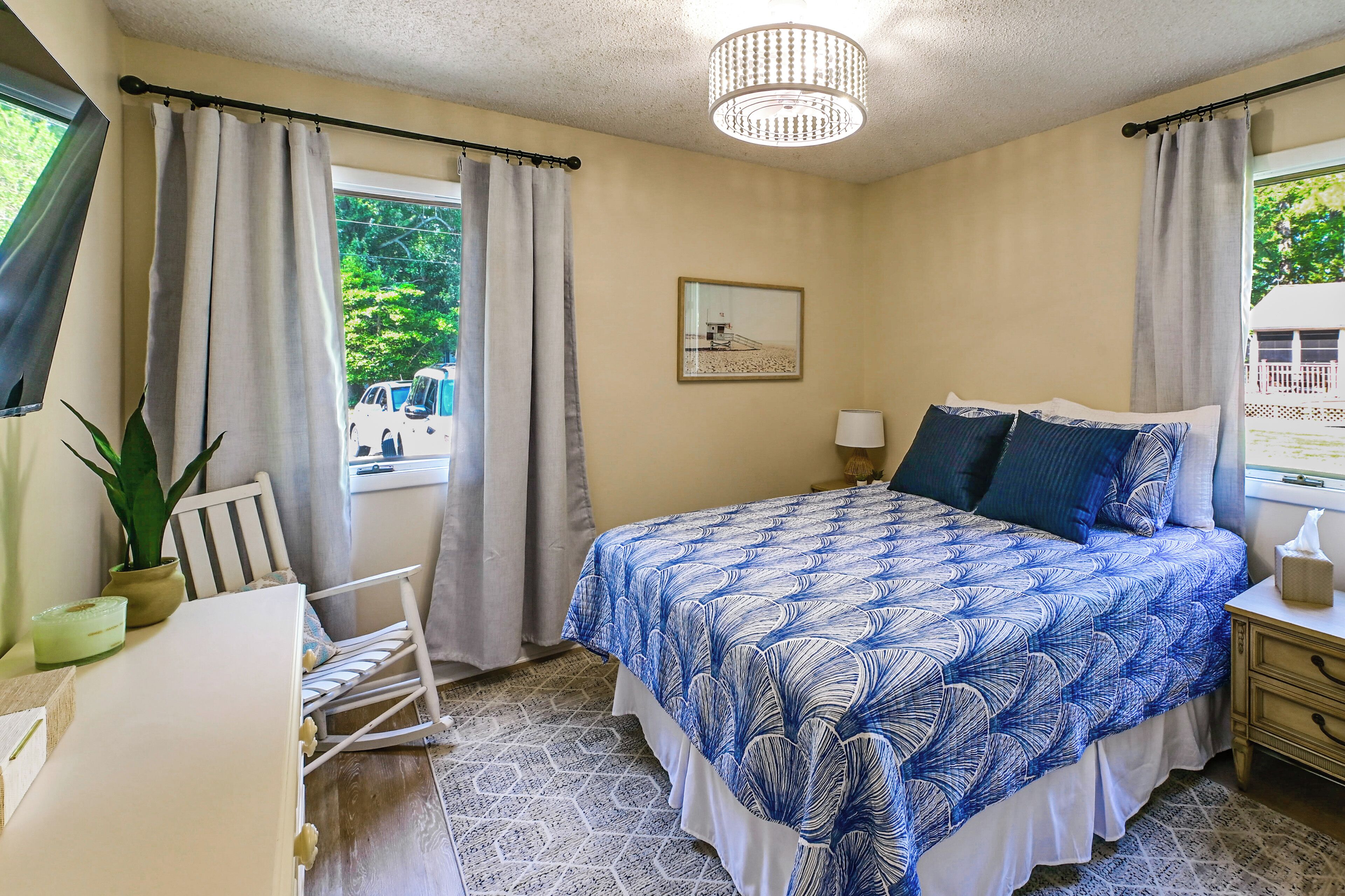 A bedroom with a bed, a desk, a chair, a window with curtains, a chandelier, and a framed picture on the wall.
