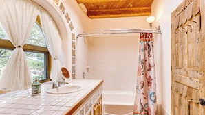 Un bany amb sostre de fusta, una finestra amb cortines blanques, un lavabo amb una ampolla decorativa, una cortina de bany amb estampats d'animals i una porta de fusta.