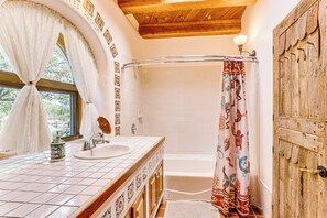 Ein Badezimmer mit Holzboden, einem Fenster mit weißen Vorhängen, einem Waschbecken mit dekorativer Flasche, einer Duschtrennwand mit Tiermustern und einer Holztür.