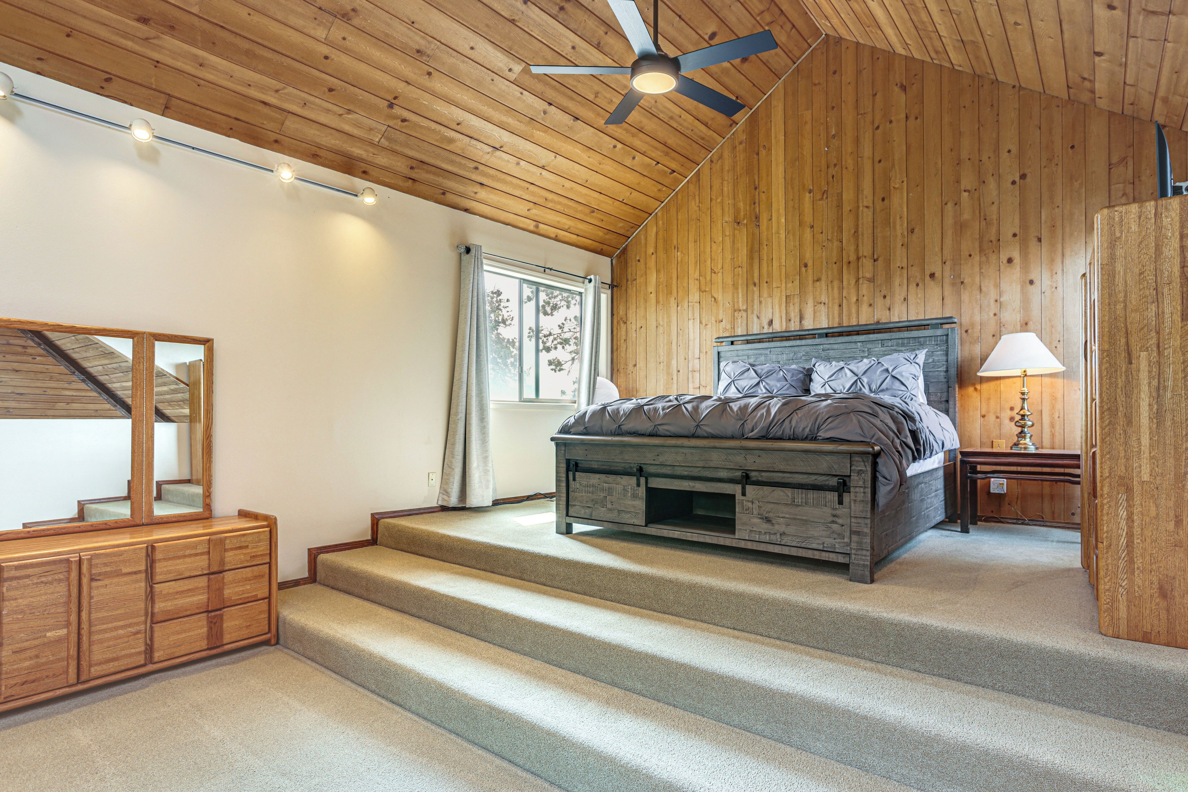 Une chambre avec un plafond en bois, un lit avec une tête de lit, une commode, une fenêtre et un ventilateur de plafond.